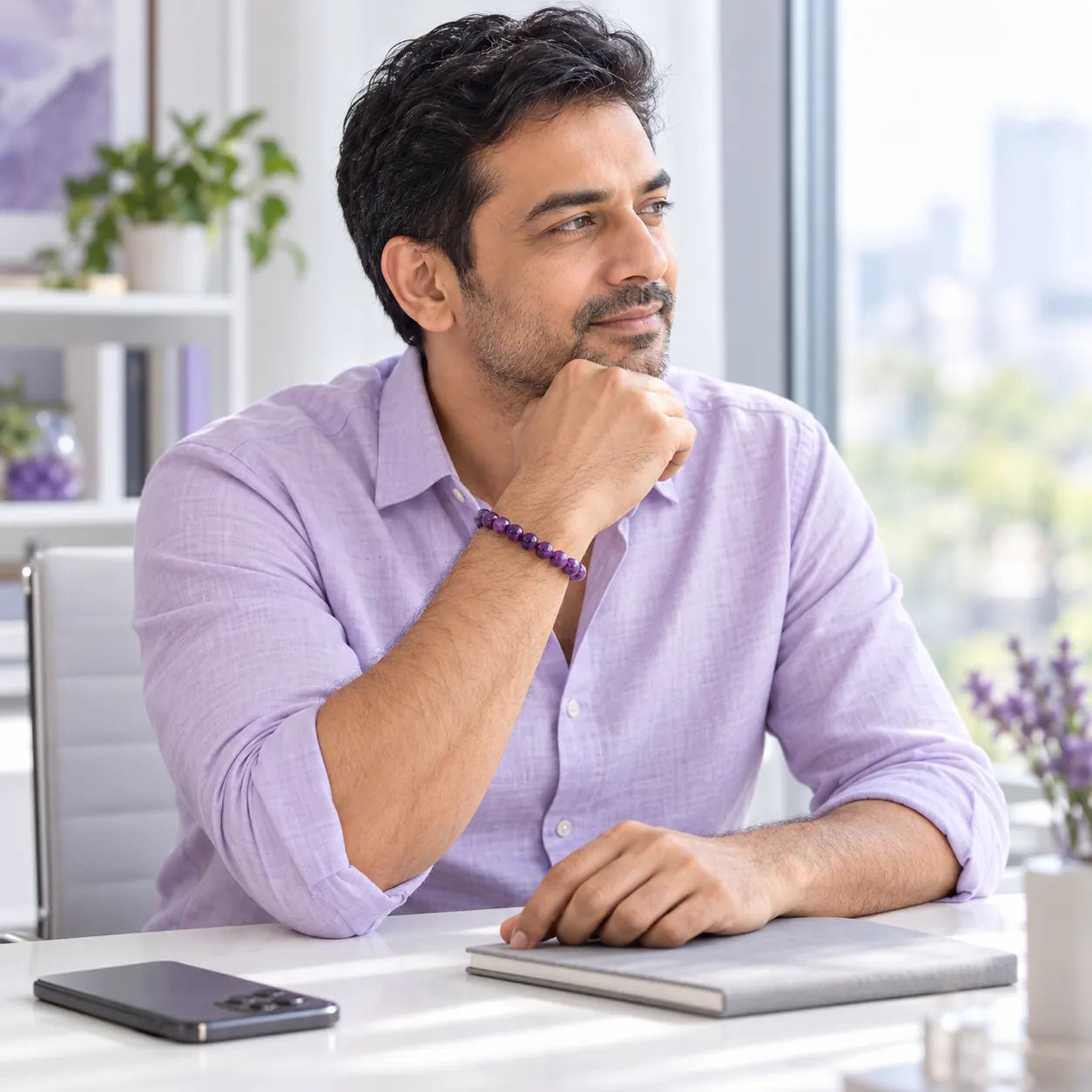 An Indian man wearing a purple amethyst bracelet takes a thoughtful pause beside a bright window, with his notebook closed and phone face-down