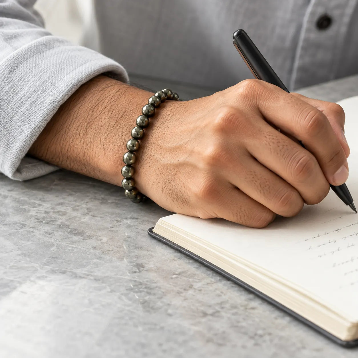 A hand wearing a metallic Pyrite bead bracelet writes in a notebook on a pale grey desk